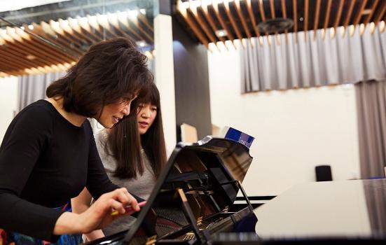 Two Asian woman looking at a musical score over a piano. The photo is shot from the side. The woman closest to the camera is wearing a black top, she has short brown hair with a fringe, and is holding a pencil. The woman next to her has long, dark brown hair with a fringe, and is wearing a grey top.