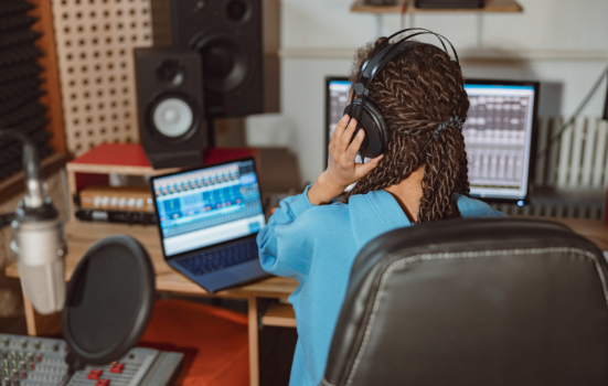 A young person sits at a music production desk. She is holding headphones with her back to the camera